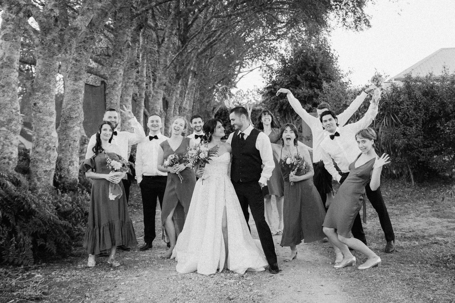 Black and white wedding photo of bride, groom, and bridal party celebrating joyfully among tall trees.