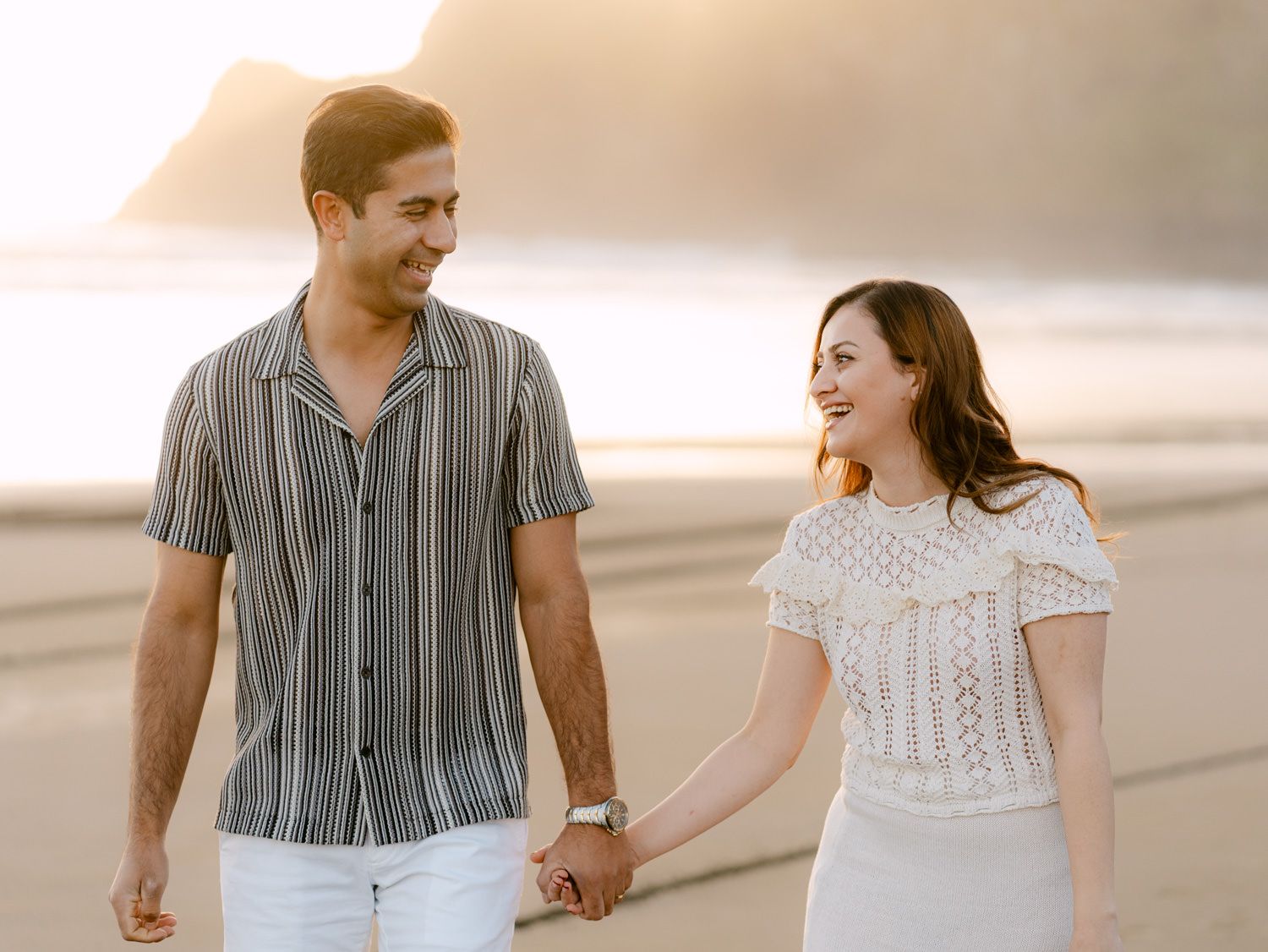 Couple holding hands and smiling at each other while walking on a sandy beach at golden hour sunset.