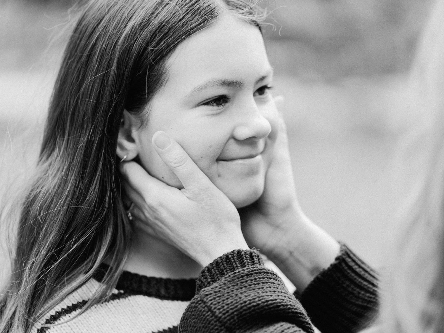 Black and white photo of a smiling girl having her face gently held by caring hands.