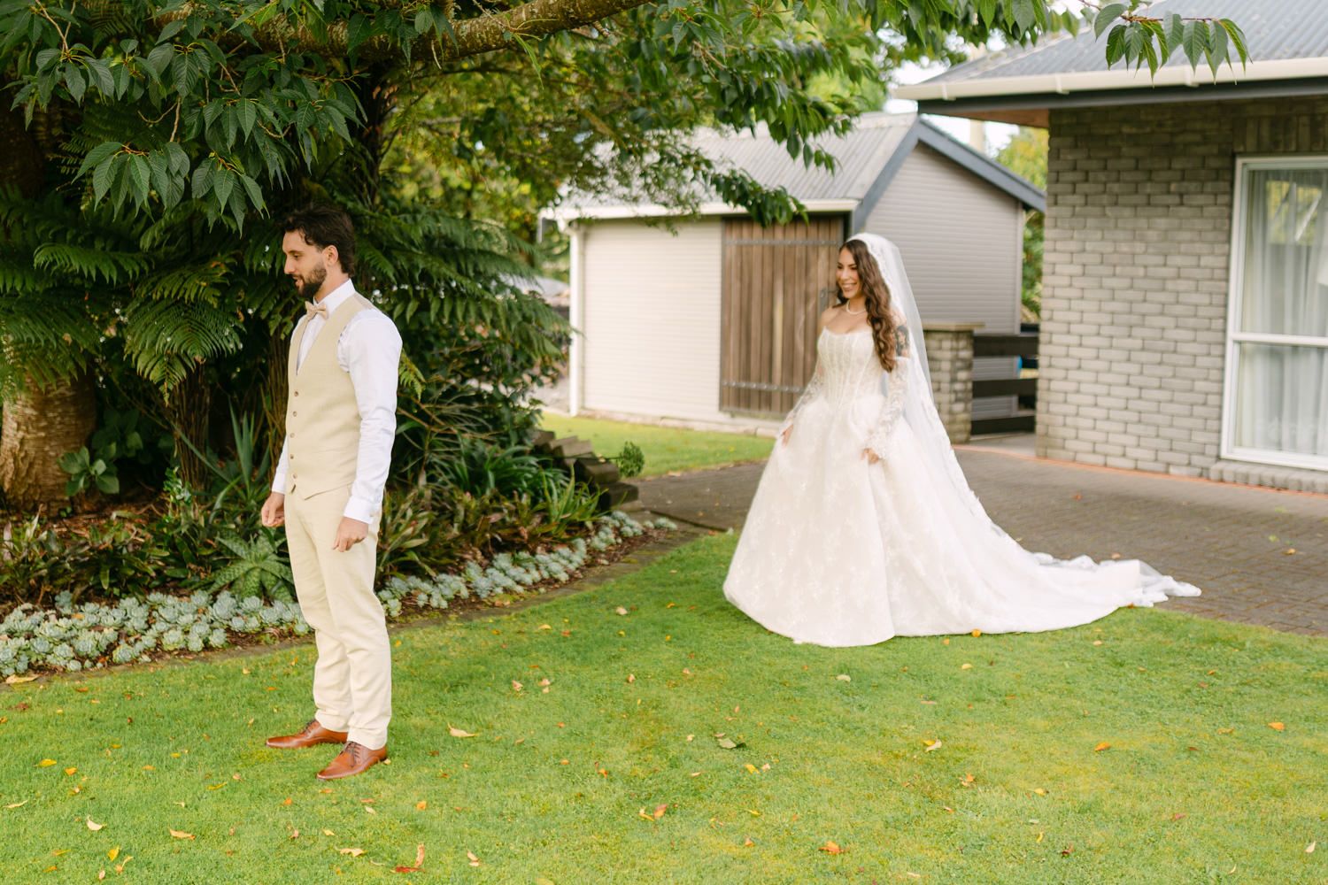 A bride in a white gown and groom in a beige suit stand on a lush green lawn outside a house.