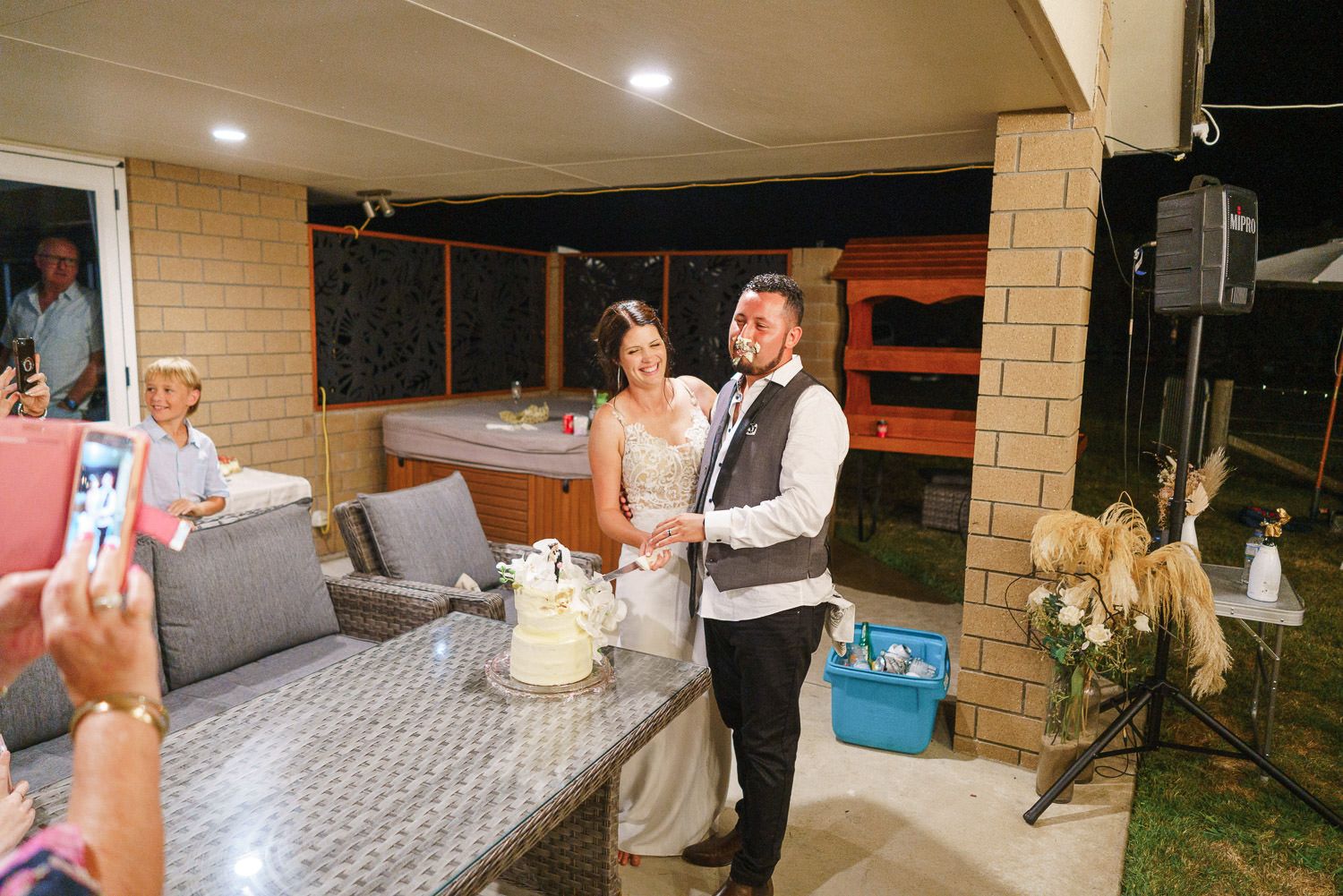 Bride and groom cutting wedding cake at outdoor reception under patio lights at night.