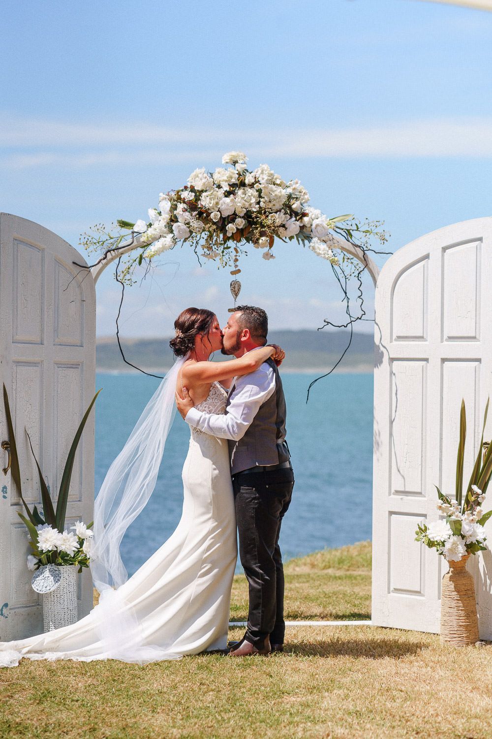 Bride and groom sharing first kiss under white floral arch by ocean.