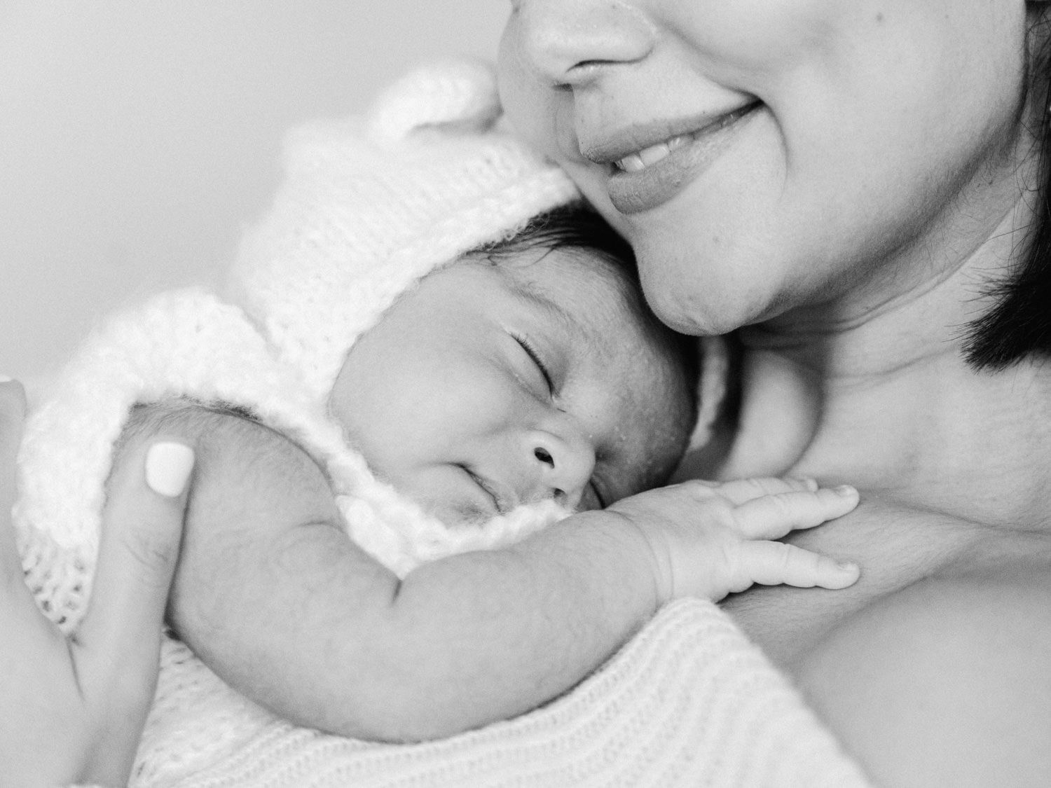 Black and white photo of mother gently kissing sleeping newborn baby in white knit bonnet.