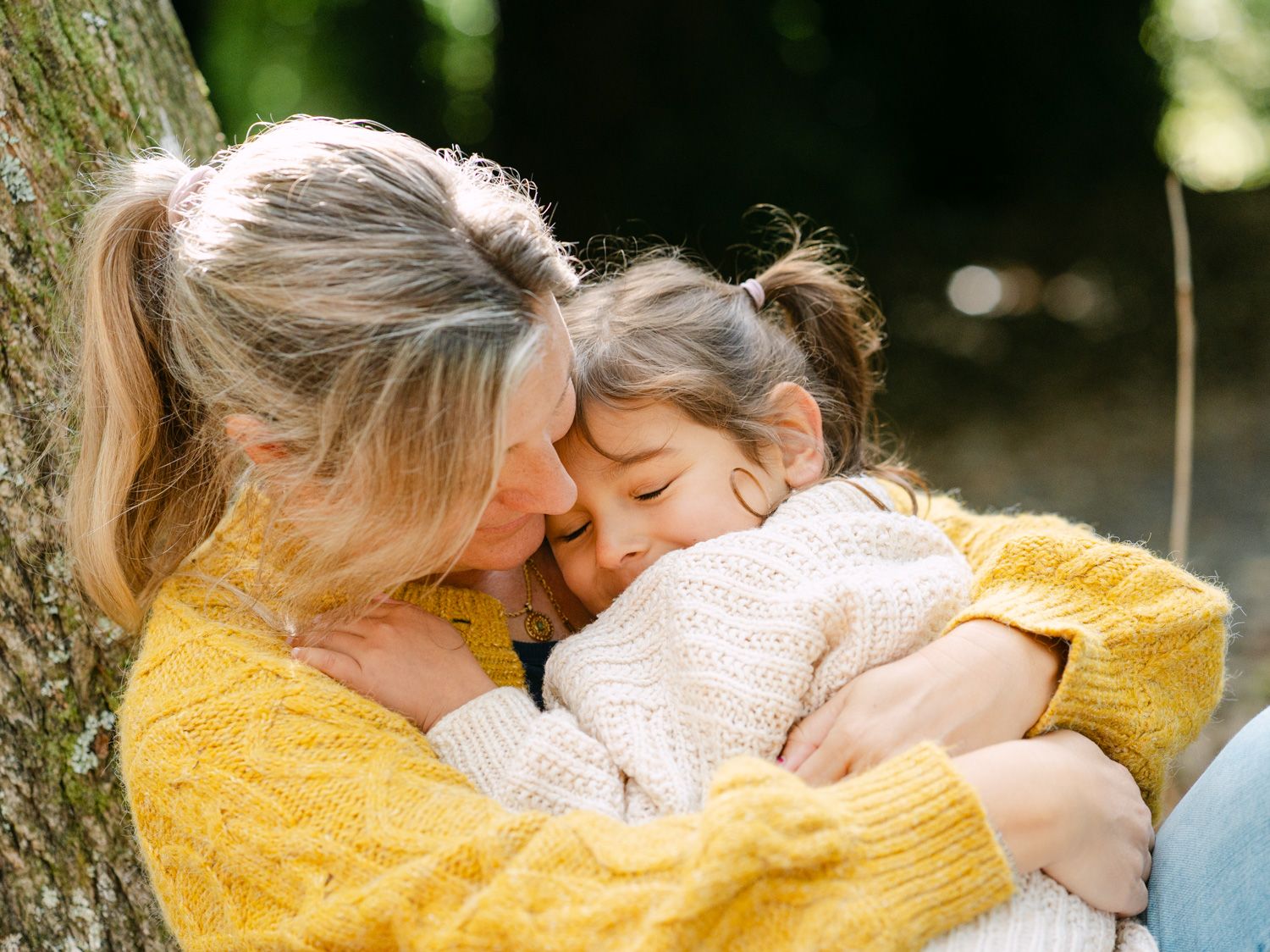 A mother in a yellow sweater tenderly embraces her young daughter outdoors near a tree.