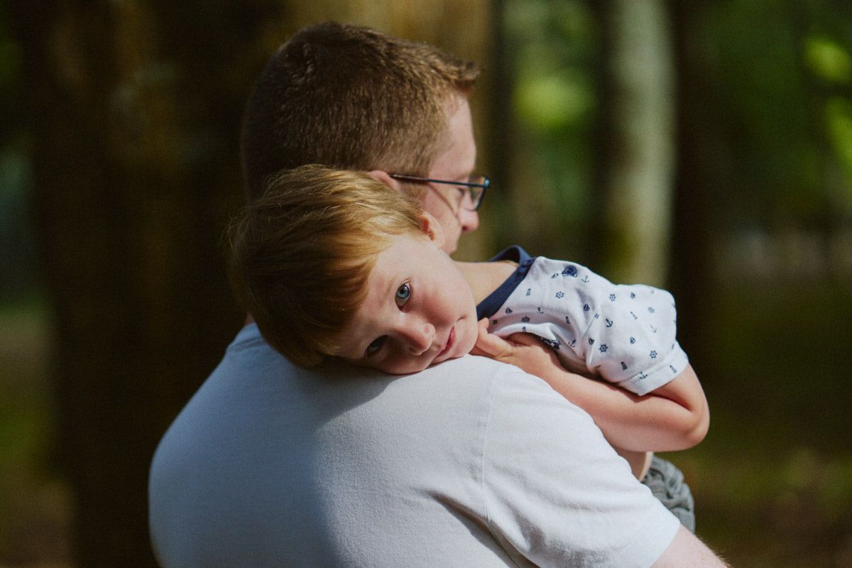 A father tenderly holds his sleepy young child against his shoulder outdoors in a sunlit forest setting.