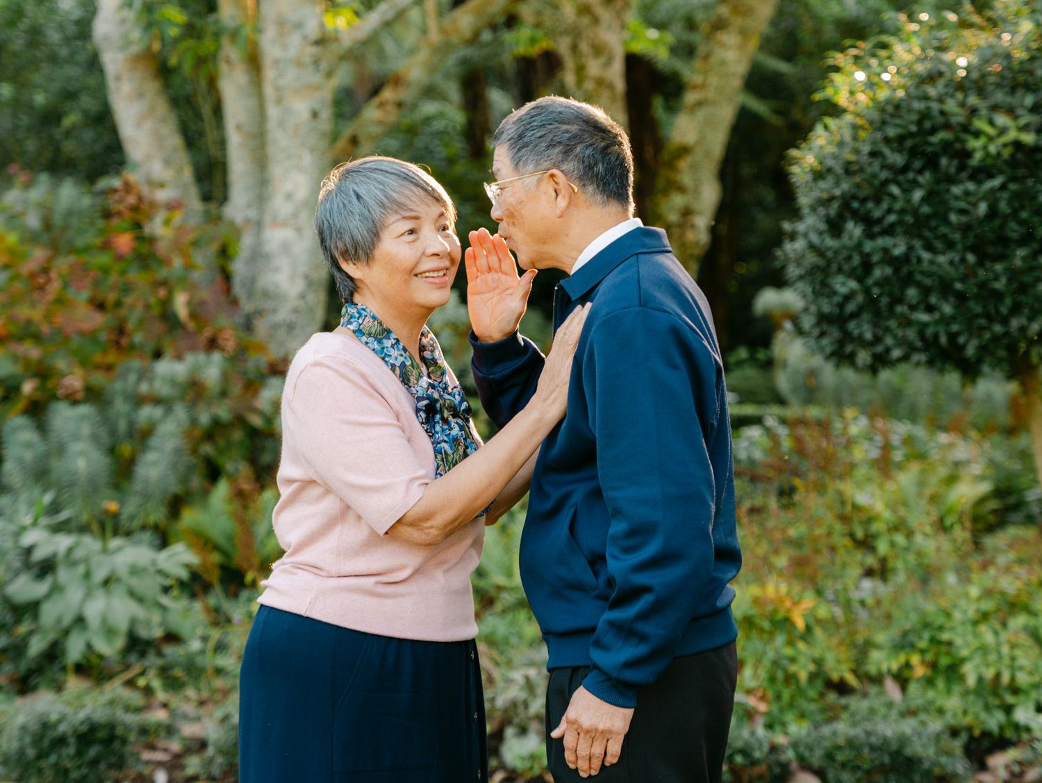 Elderly Asian couple sharing a tender moment in a lush green garden, smiling and holding hands.
