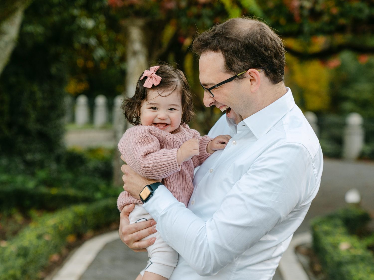 A father in a white shirt holds his smiling baby girl in a pink outfit in a lush garden setting.