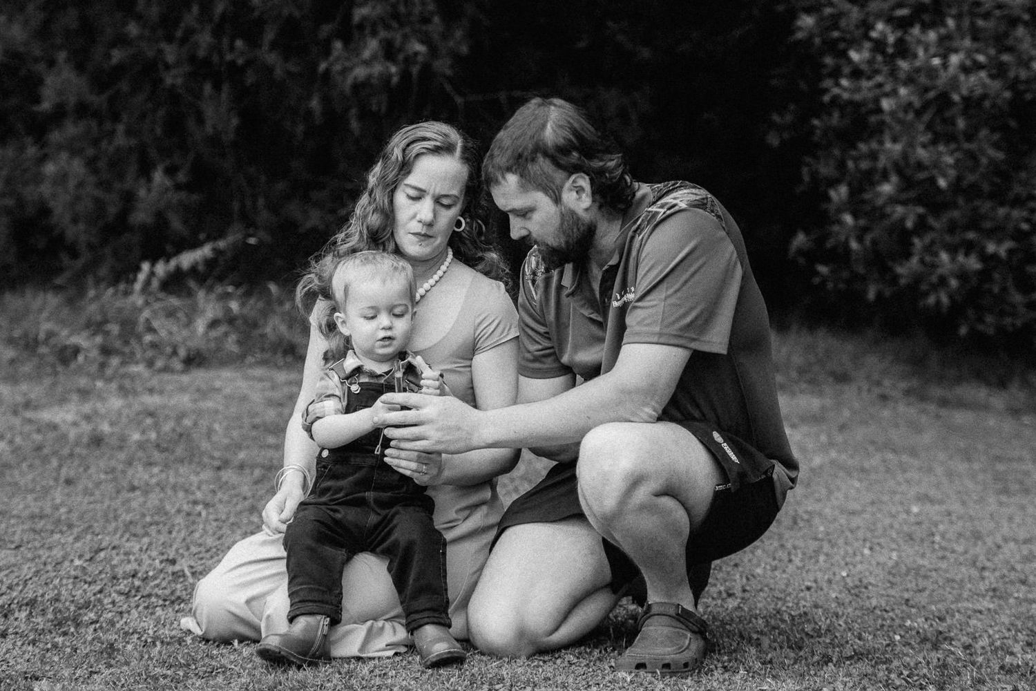 Black and white photo of a young family with a toddler sitting on grass, parents smiling while showing child something on a phone.