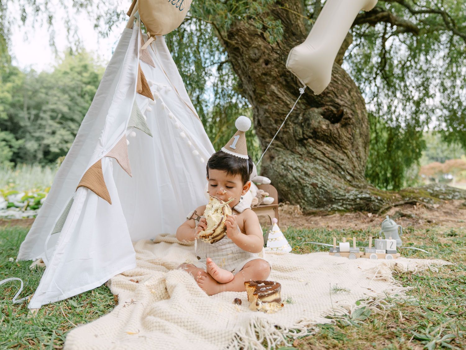 Baby in party hat enjoys cake smash outdoors near white teepee tent on blanket in garden setting.