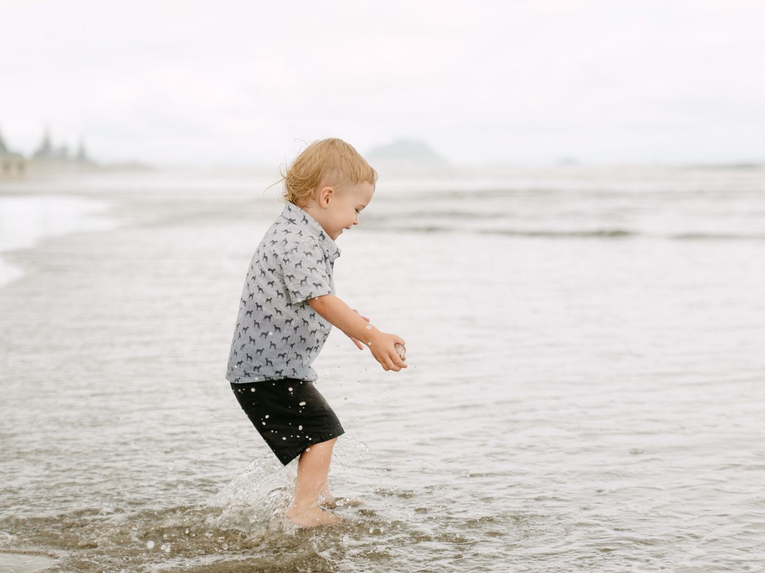 A young toddler boy splashing in shallow ocean waves on a sandy beach wearing a patterned shirt and black shorts.