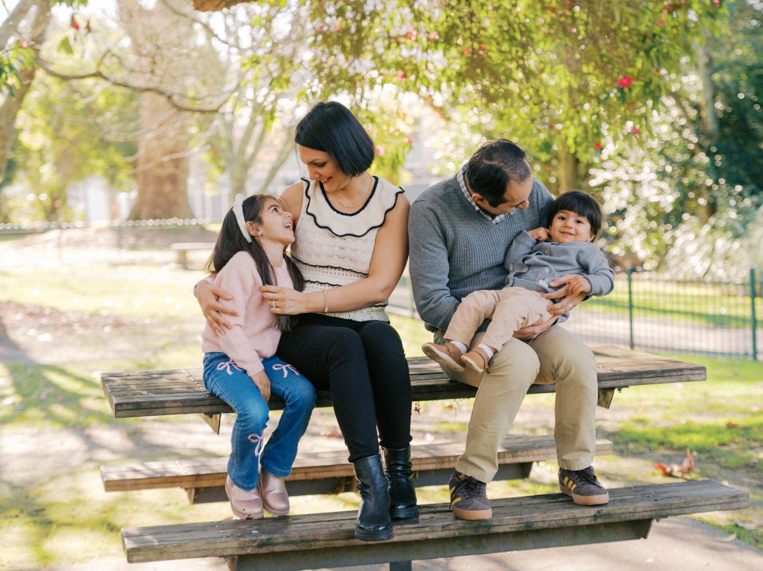 Family of four sitting on a wooden park bench surrounded by green trees on a sunny day.