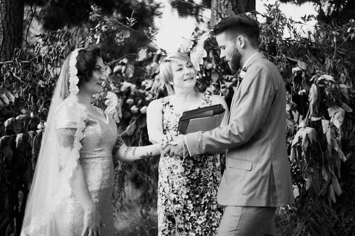 Black and white wedding ceremony photo of bride and groom holding hands before officiant outdoors.