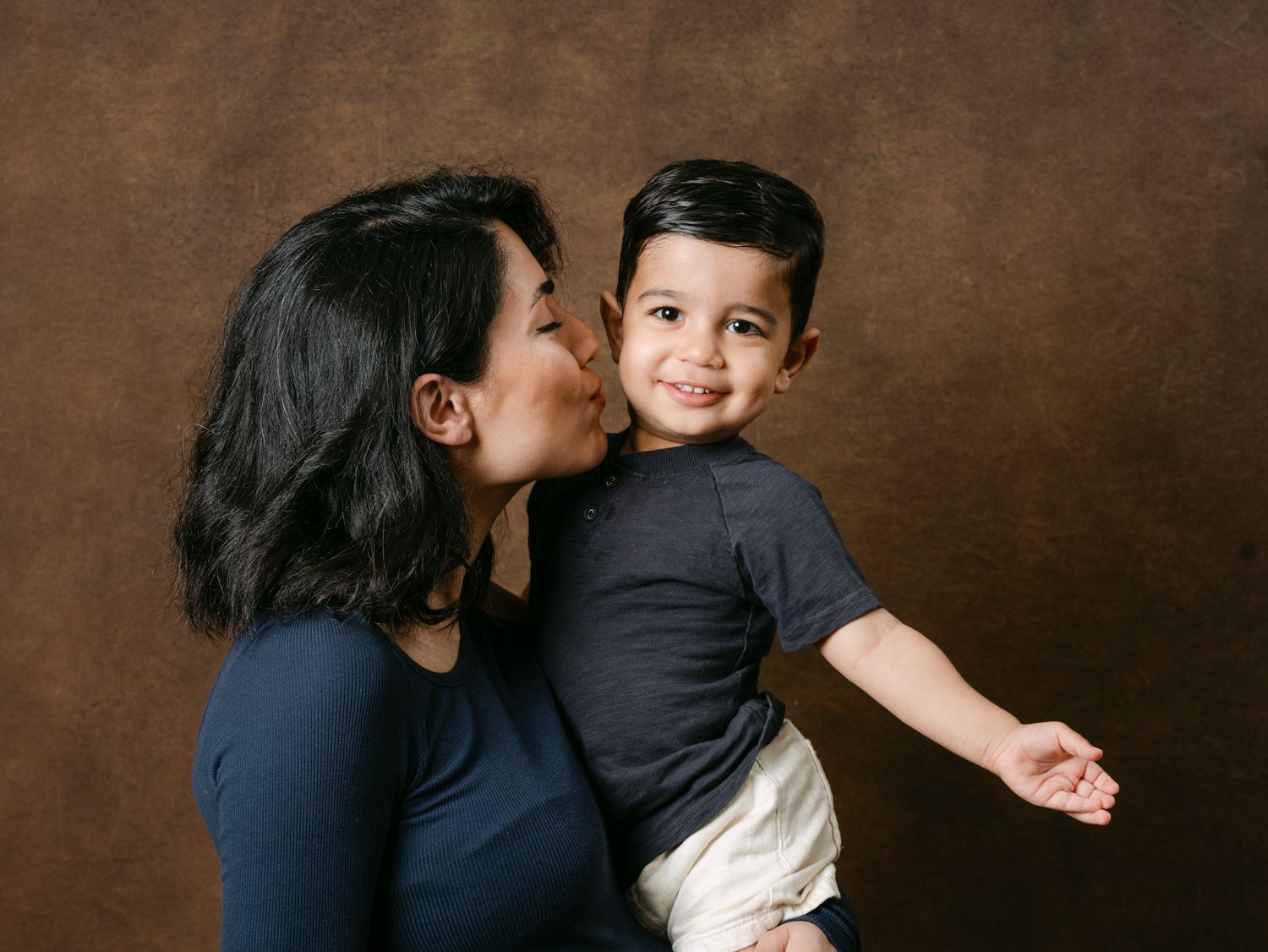 A woman in a navy top kisses a smiling toddler boy she holds against a brown studio backdrop.