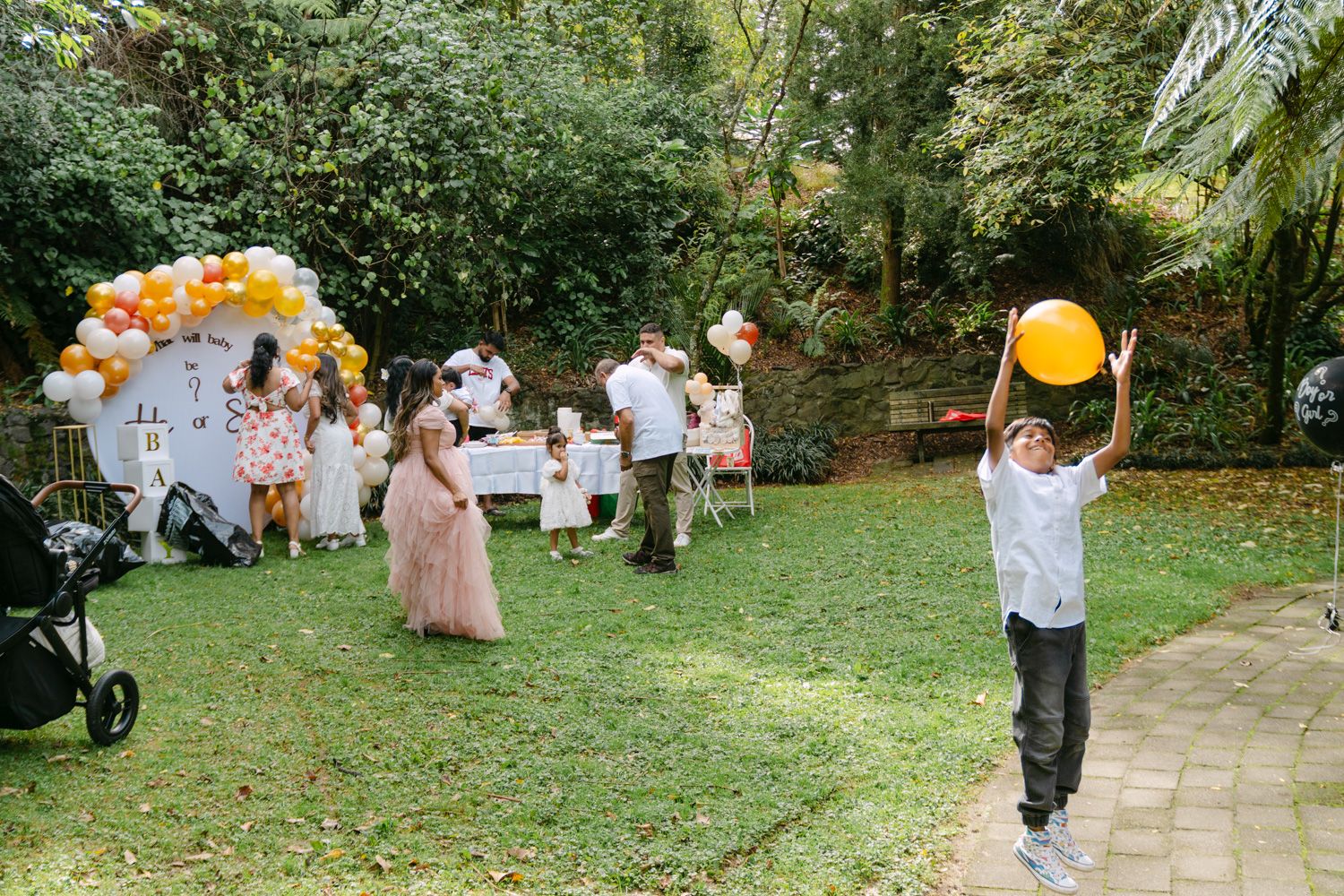 Children playing with yellow balloons at an outdoor birthday party with balloon arch decorations in a garden.