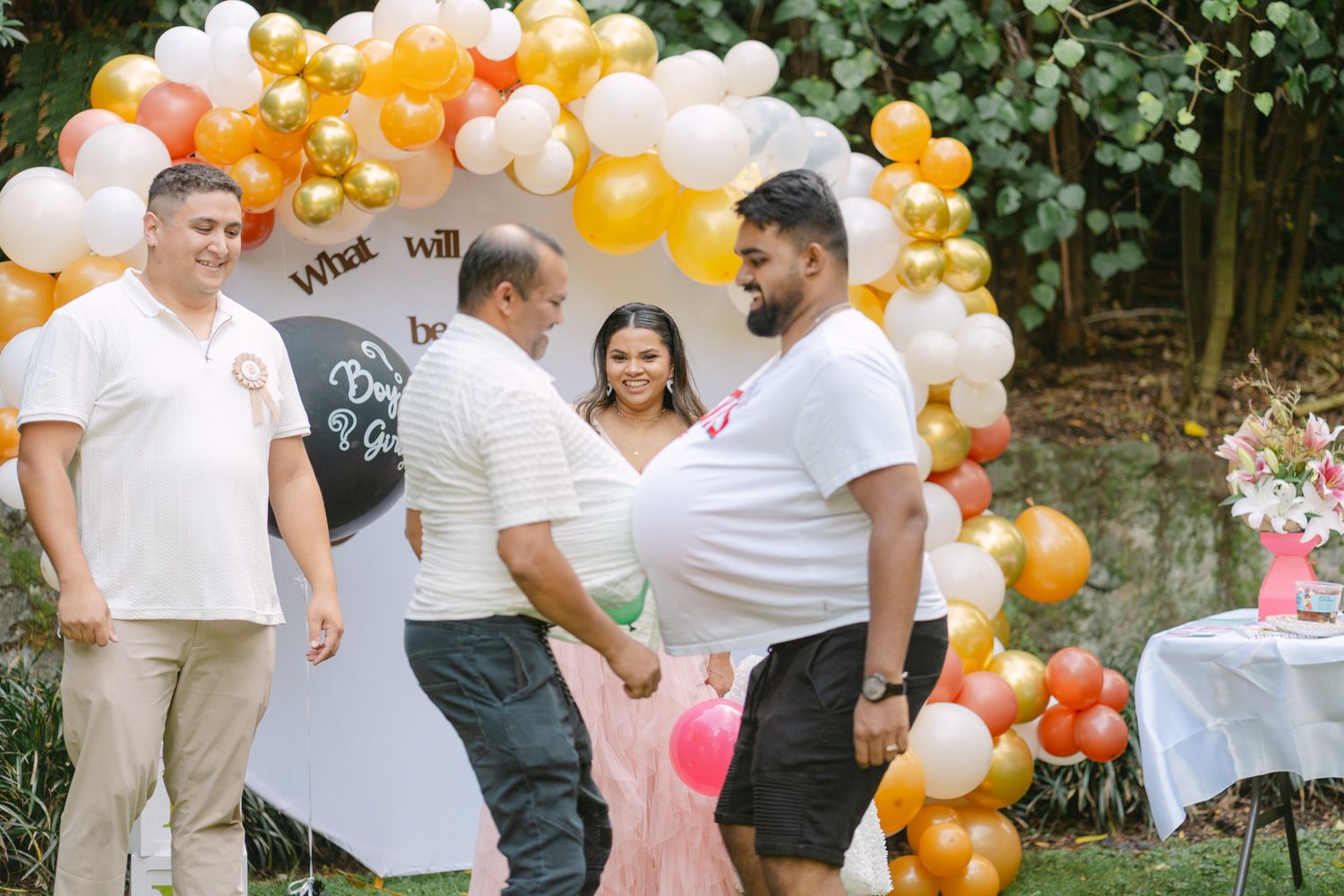 Outdoor gender reveal party with balloon arch in gold, white, and orange colors with guests gathered around.