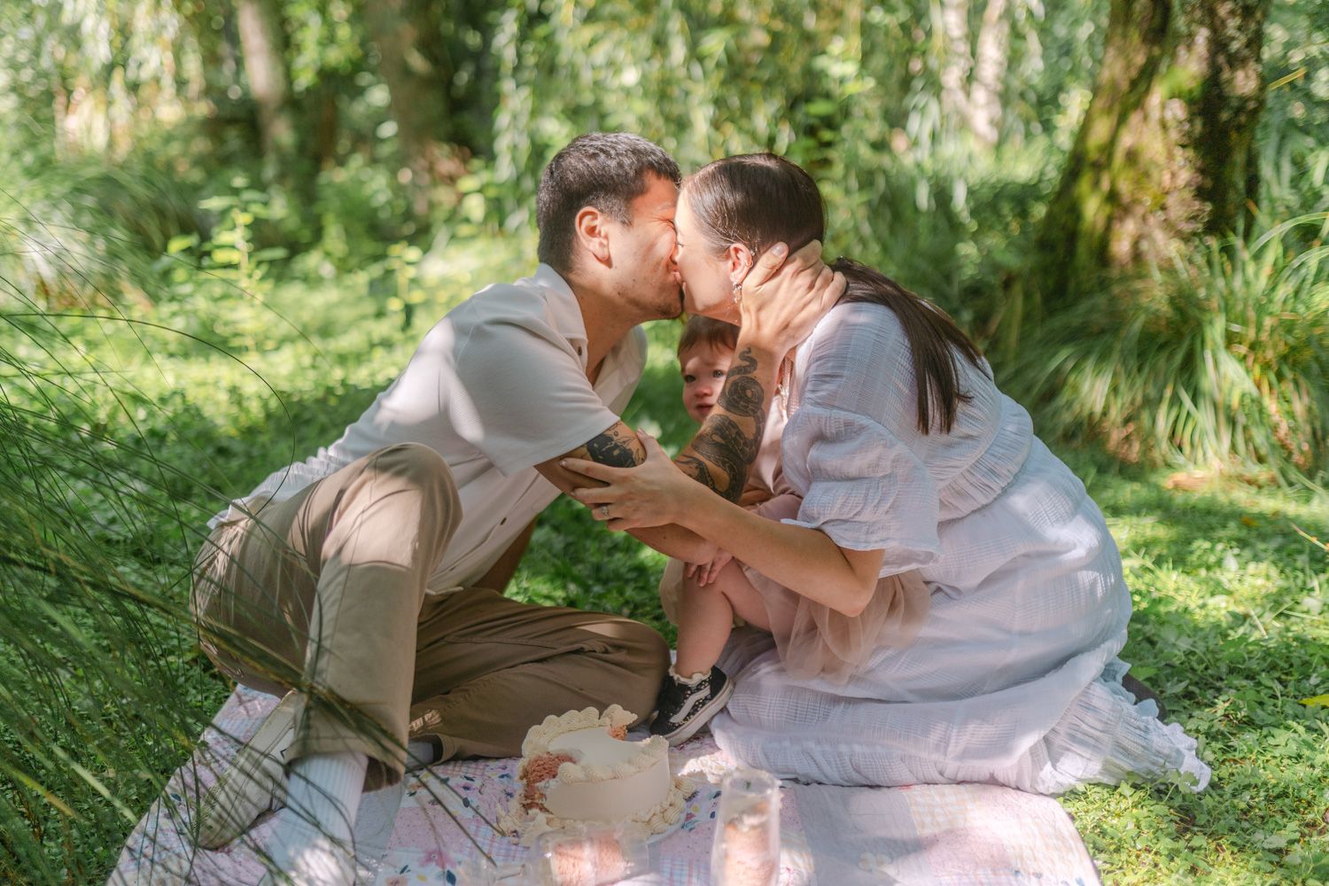 A couple shares a tender kiss while sitting on a picnic blanket in a lush green park with their child.