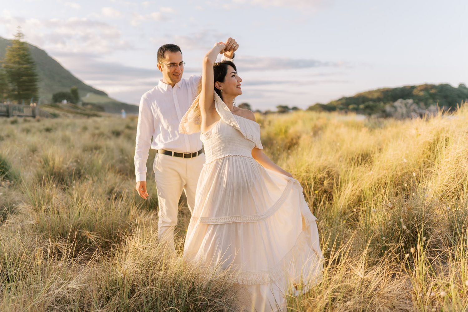 Couple dancing in golden field at sunset, woman in white tiered dress twirling, man in white shirt smiling outdoors.