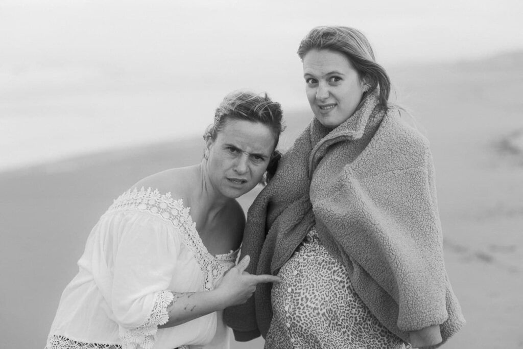 Black and white beach photo of two women posing together, one kneeling, wrapped in a blanket on a sandy shore.