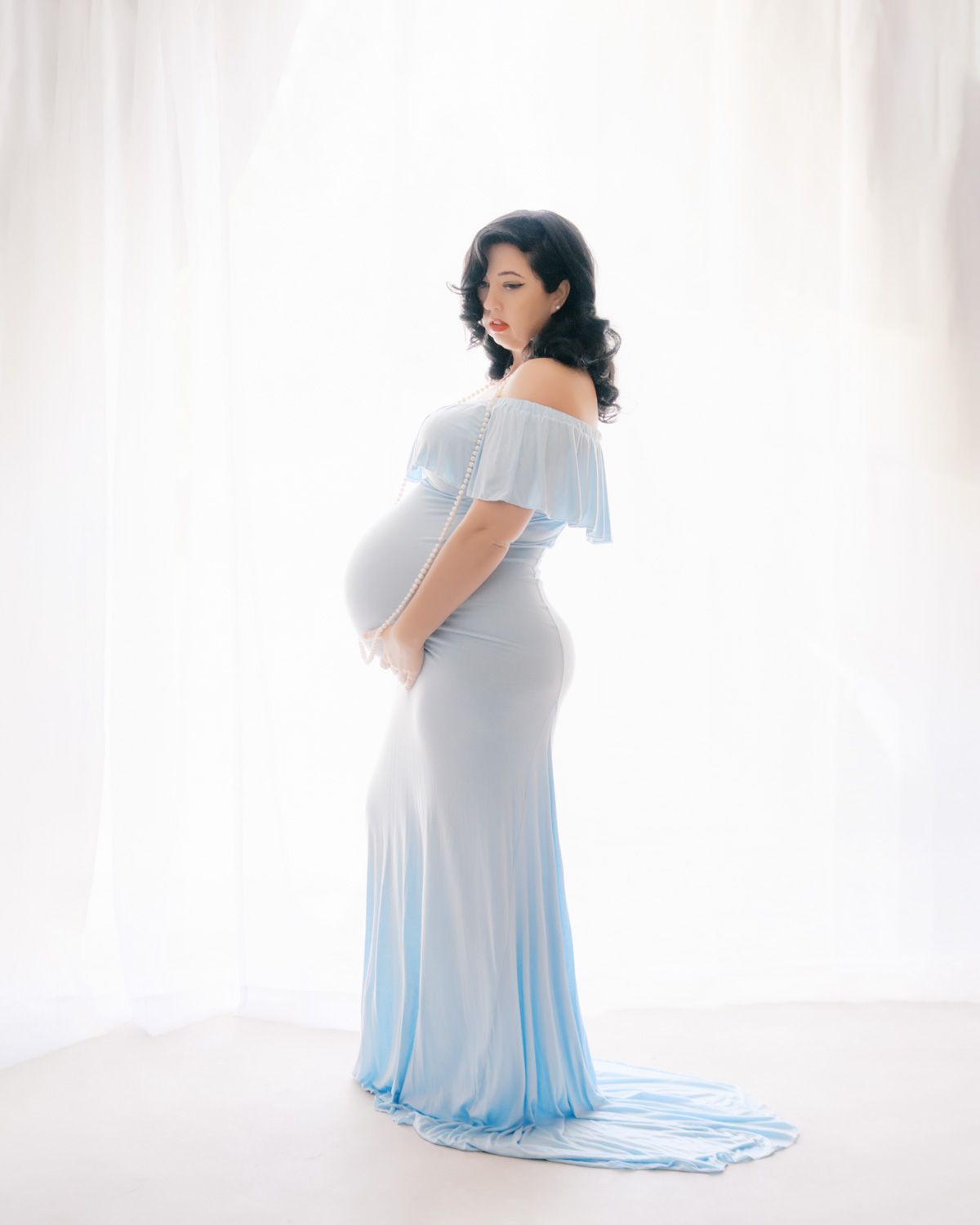 Pregnant woman in elegant light blue off-shoulder maternity gown posing gracefully against white curtains.