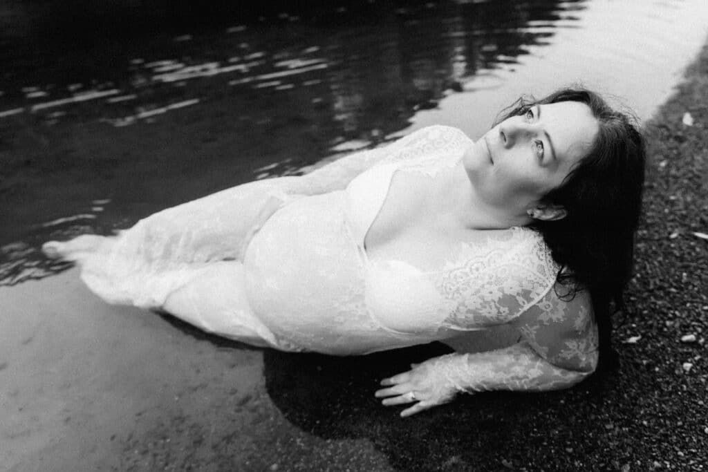 Black and white photo of a woman in a white lace dress lying in shallow water, looking upward peacefully.