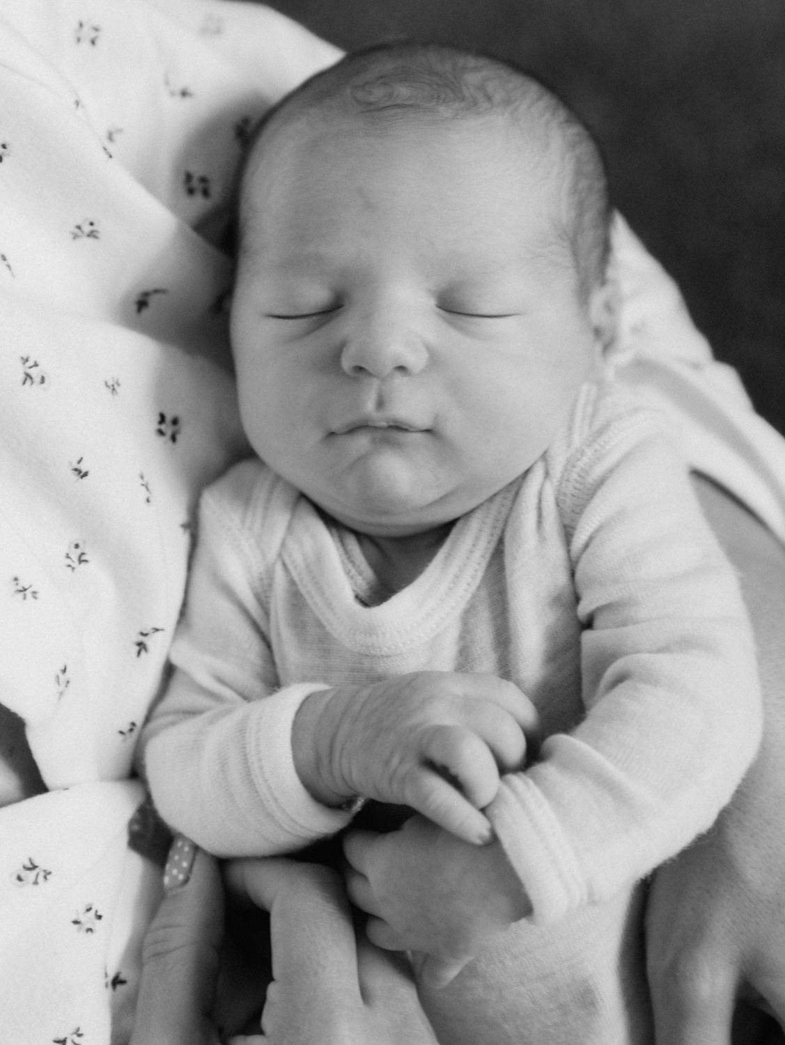 Black and white photo of a sleeping newborn baby in a onesie, peacefully resting with tiny hands visible.