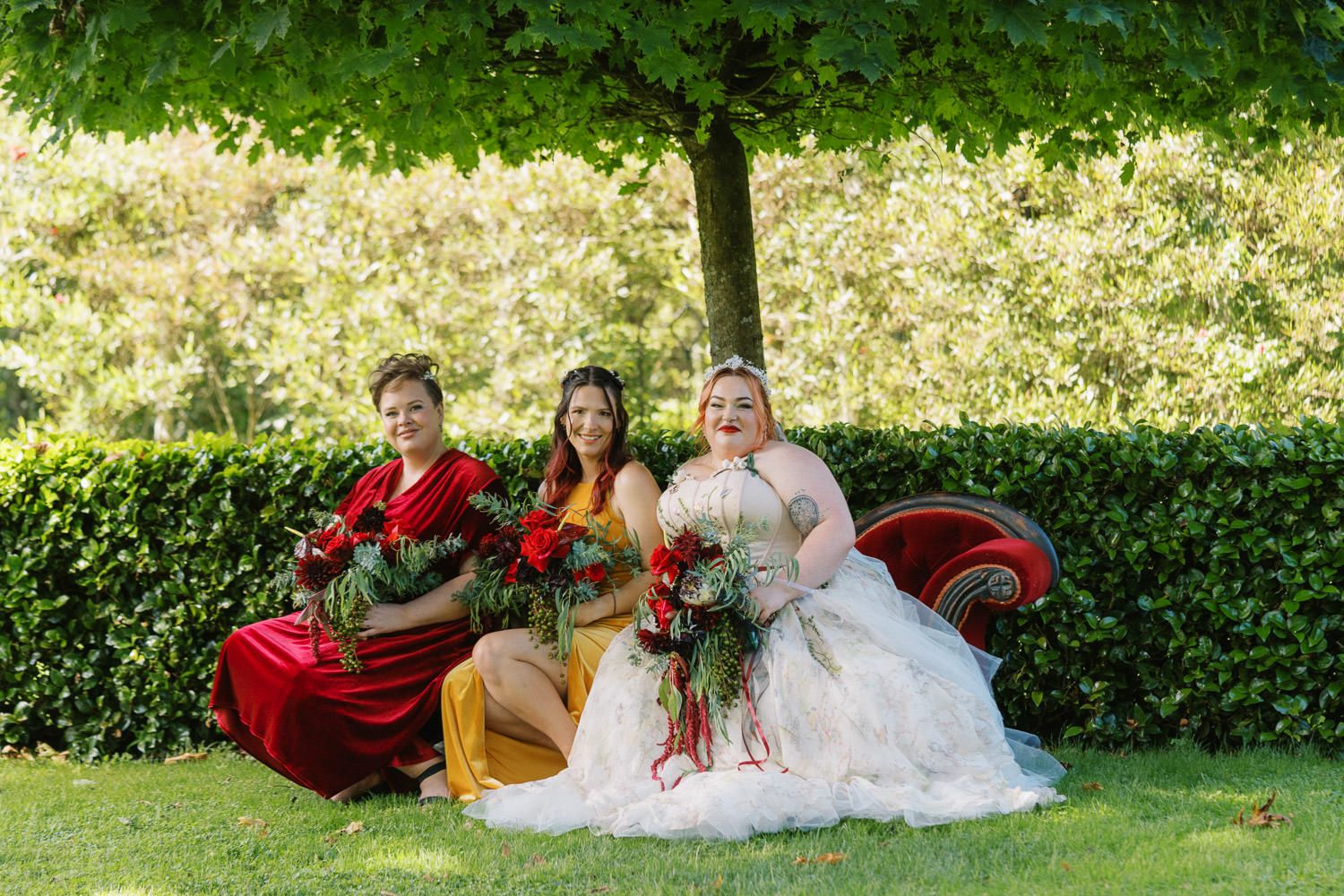 Bride in white gown poses with two bridesmaids in red and yellow dresses holding bouquets outdoors.