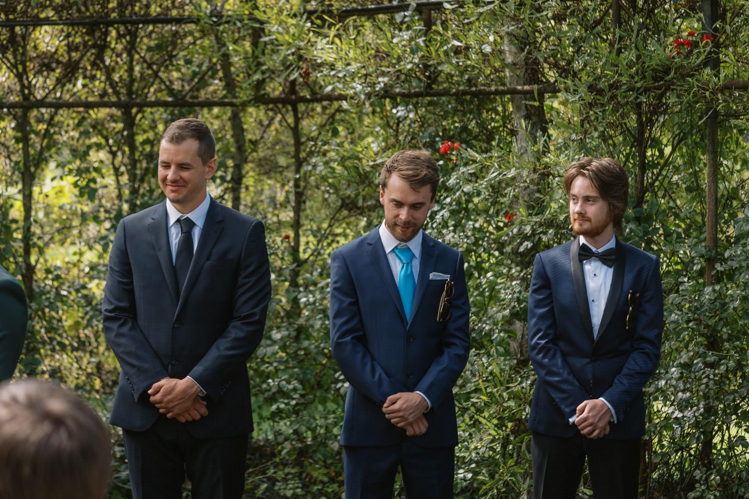 Three men in suits stand outdoors at a wedding ceremony, surrounded by lush green foliage and a floral arbor.