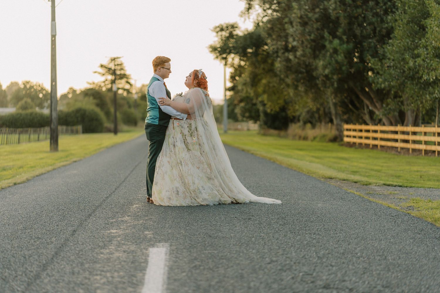 Bride and groom standing on a quiet rural road at sunset, sharing a romantic moment in wedding attire.