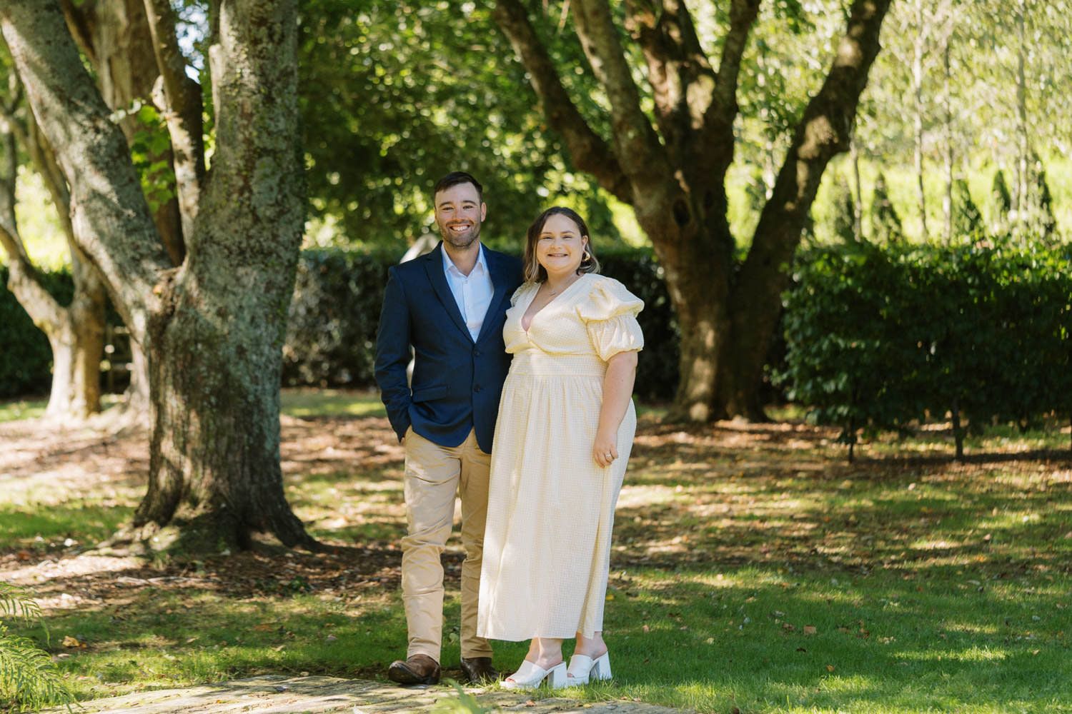 Couple posing outdoors in a lush green park, man in navy blazer and woman in yellow dress.