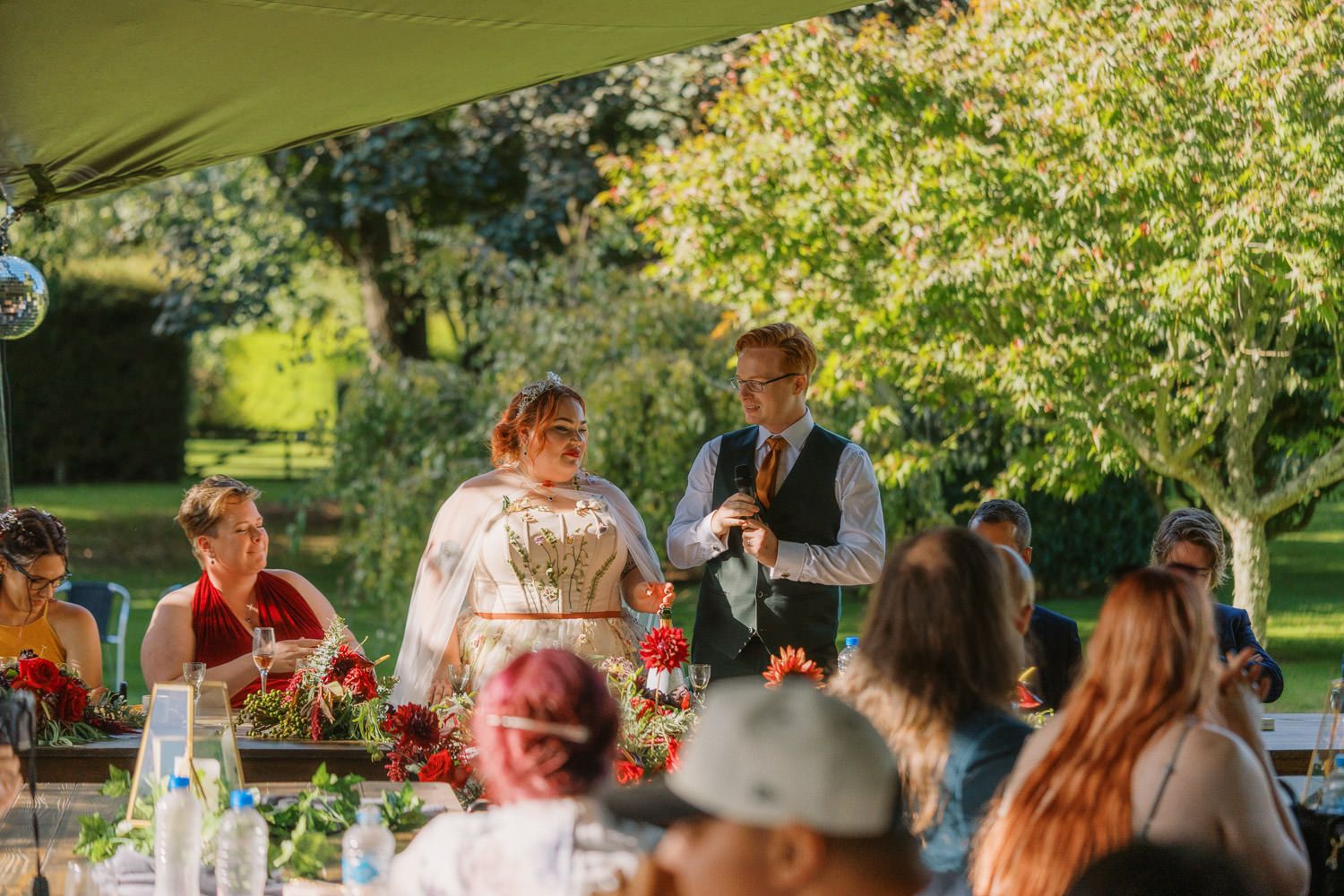 Bride and groom share a joyful moment with guests at an outdoor wedding reception under a tent surrounded by lush greenery.