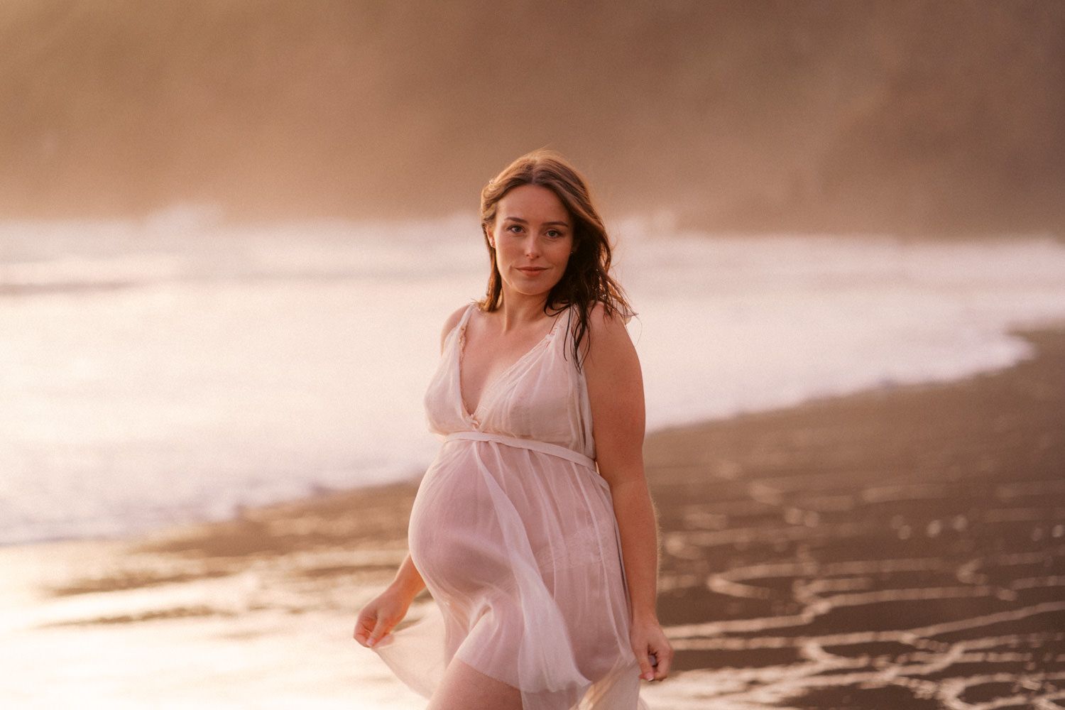 Pregnant woman in sheer white dress standing on a misty beach at golden hour with ocean waves behind her.