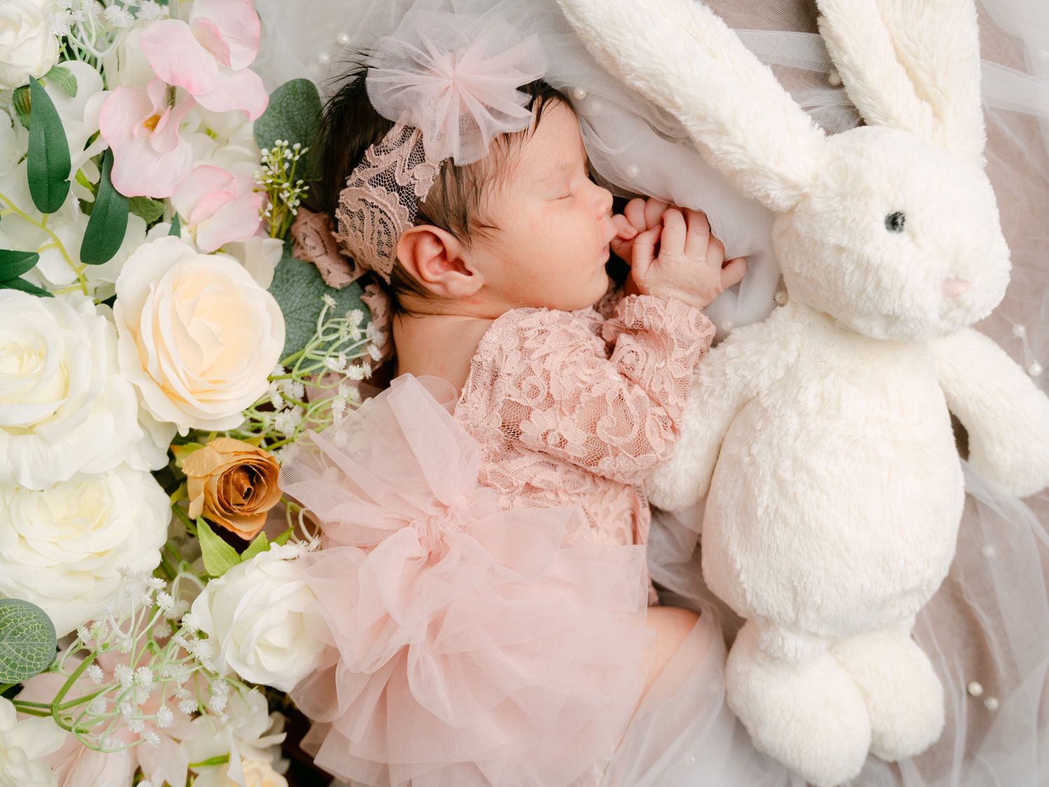 Sleeping newborn baby in pink lace dress with floral headband beside white bunny stuffed animal and fresh flowers.