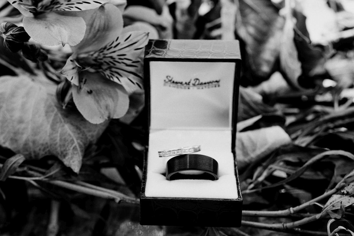 Black and white photo of a mens wedding band in a jewelry box surrounded by flowers.