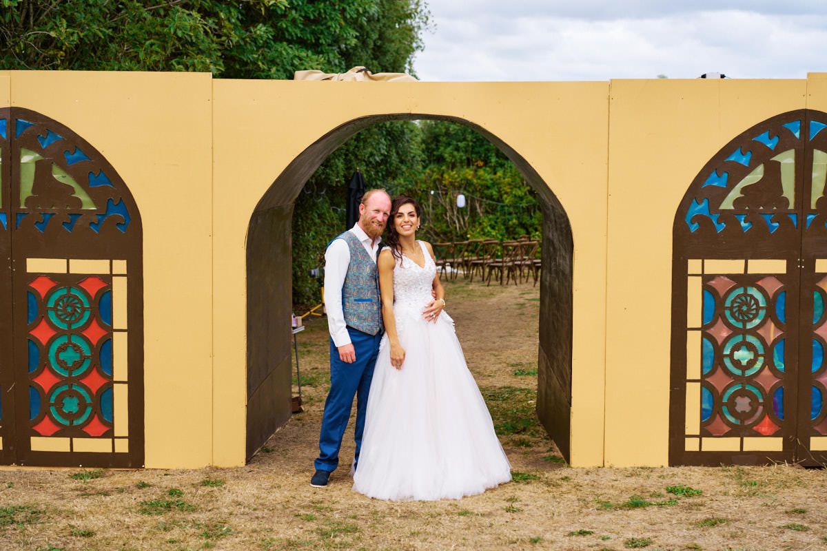 Bride and groom posing under a decorative yellow arch with stained glass panels at an outdoor wedding venue.
