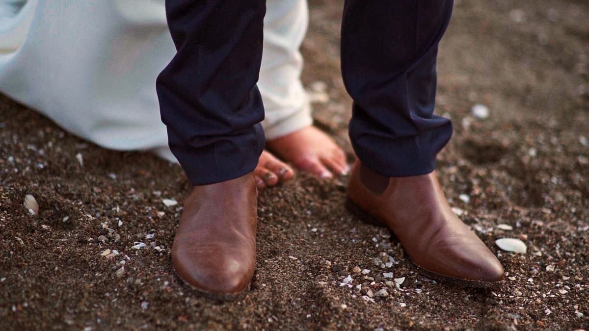 Close-up of a man wearing brown leather Chelsea boots and navy trousers standing on rocky ground.