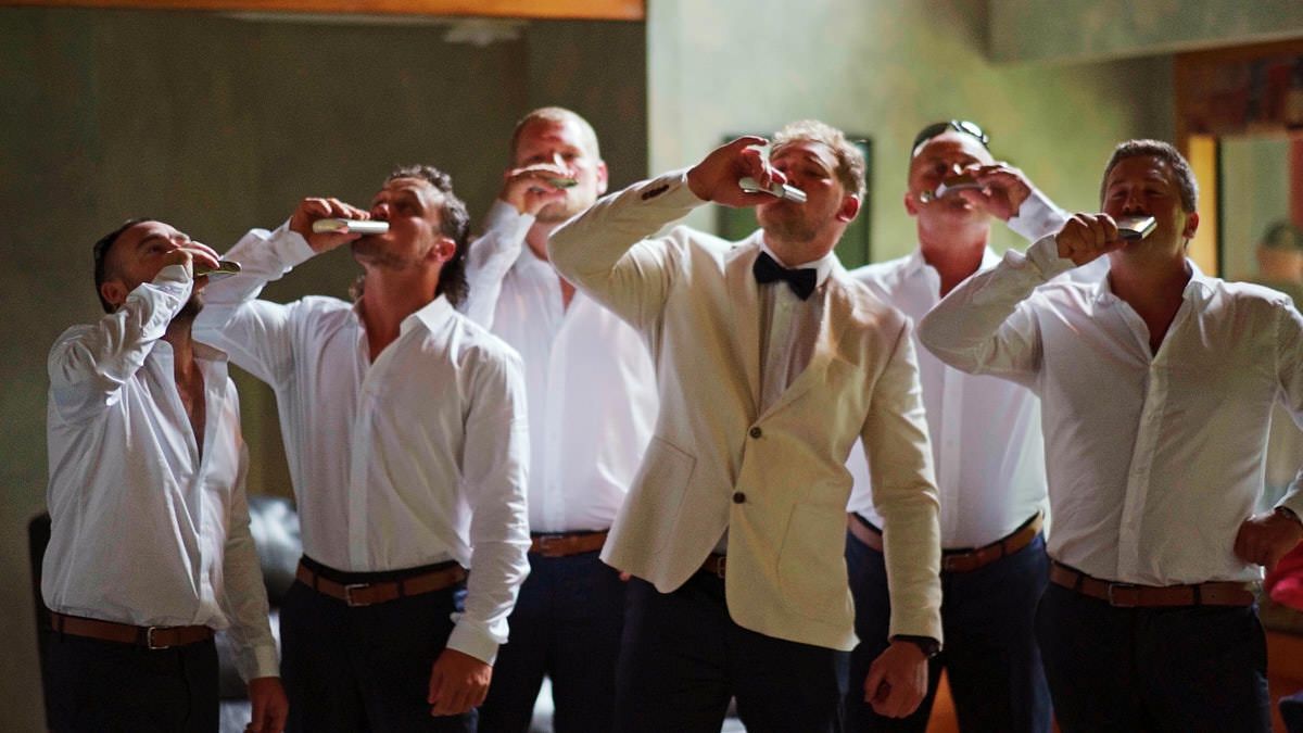 Groom in white tuxedo takes celebratory shots with groomsmen in white shirts at wedding reception.