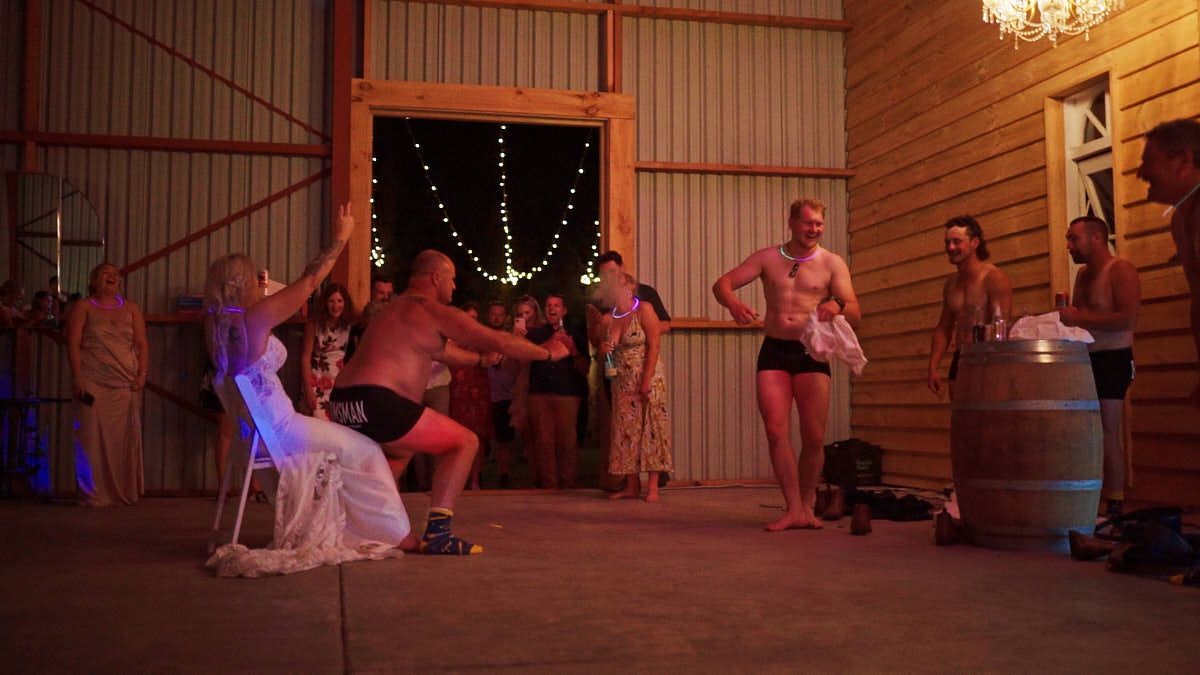Shirtless men performing at an indoor barn venue during a lively evening event.