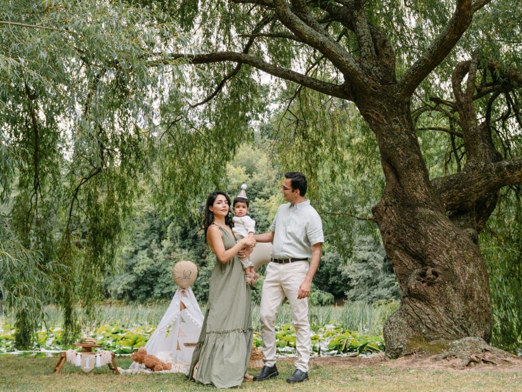 Family of three posing under a large willow tree in a lush green park during an outdoor photo session.