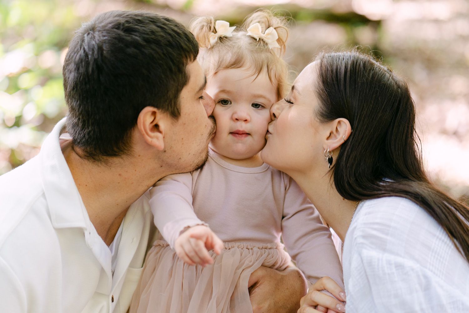 Parents kissing their adorable toddler girl on cheeks outdoors in a warm family portrait session.