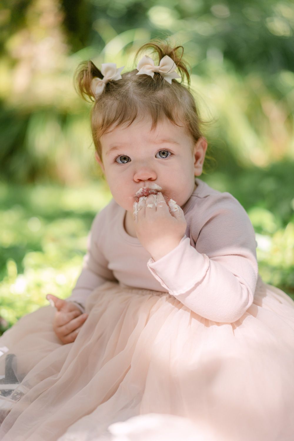 Adorable baby girl in pink tutu dress and hair bows sitting outdoors, putting hand in mouth.
