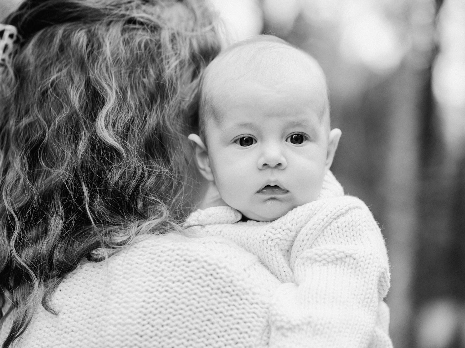 Black and white photo of a baby resting on a curly-haired woman's shoulder, looking at the camera.
