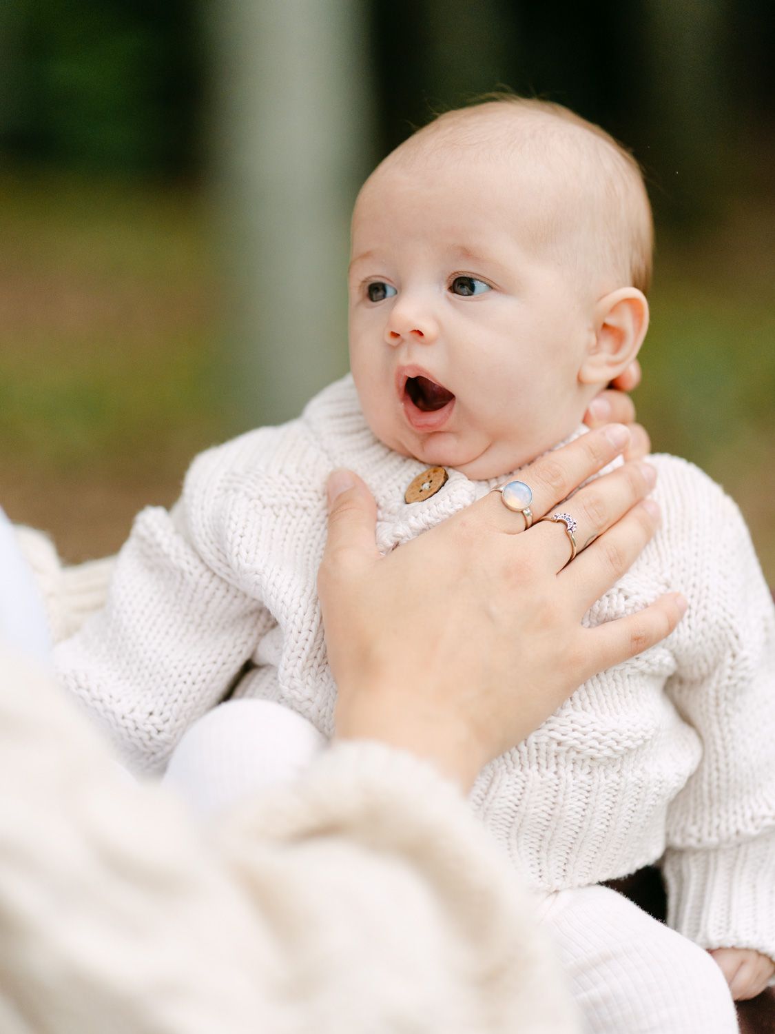 A wide-eyed baby in a white knit sweater yawns while being held by a person wearing a ring outdoors.