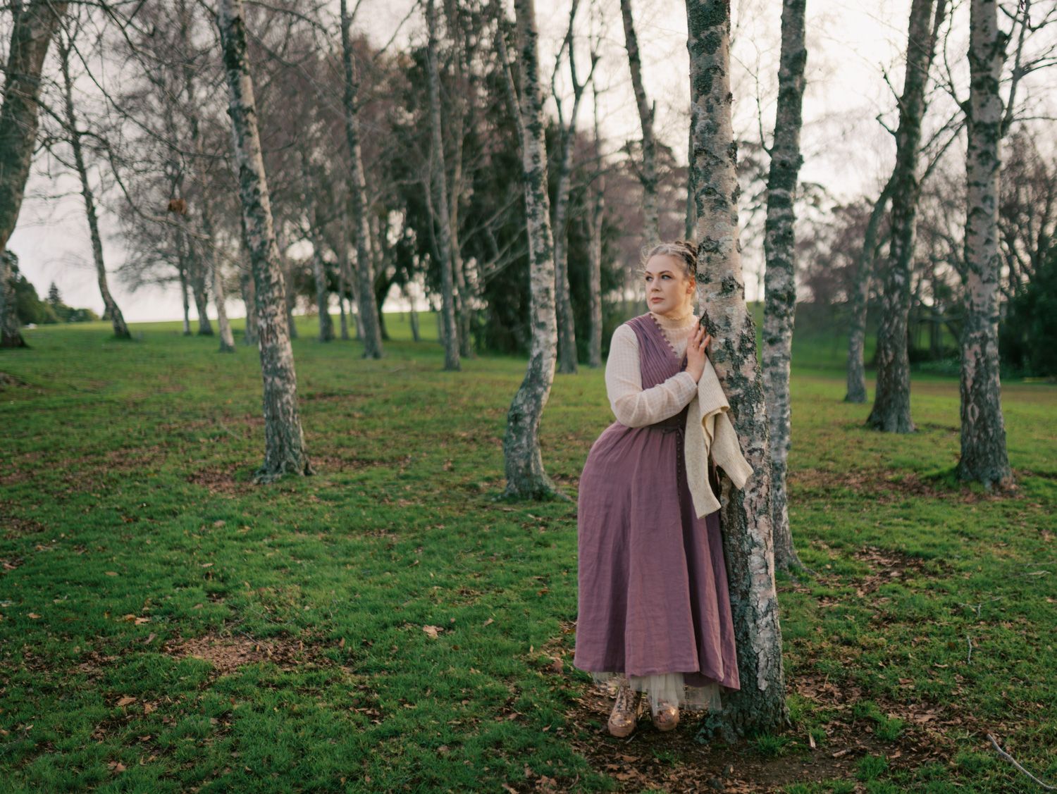Woman in purple dress leaning against birch tree in lush green woodland setting.