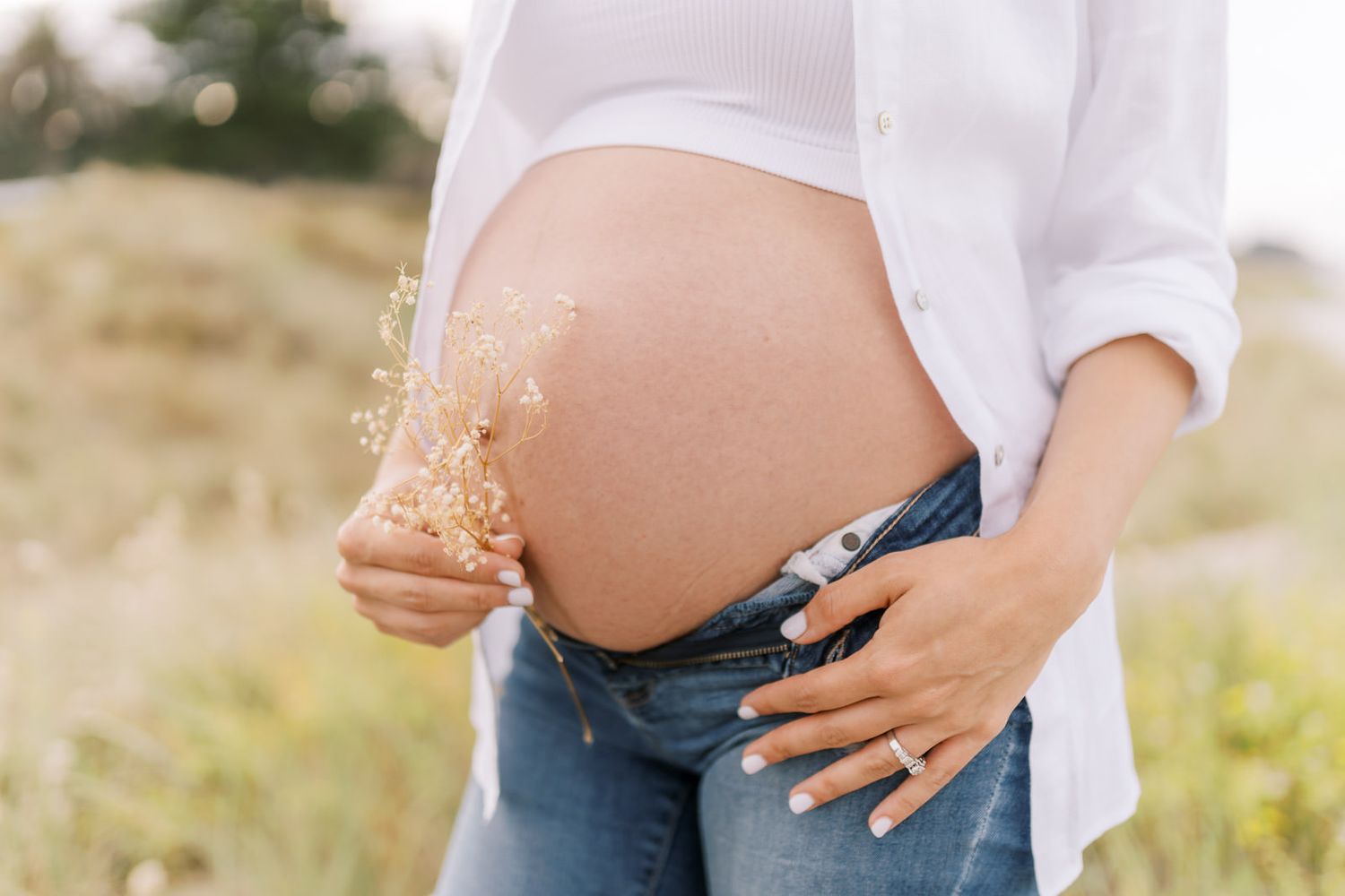 Pregnant woman in white shirt and jeans holding wildflowers, showcasing baby bump in a sunny field.