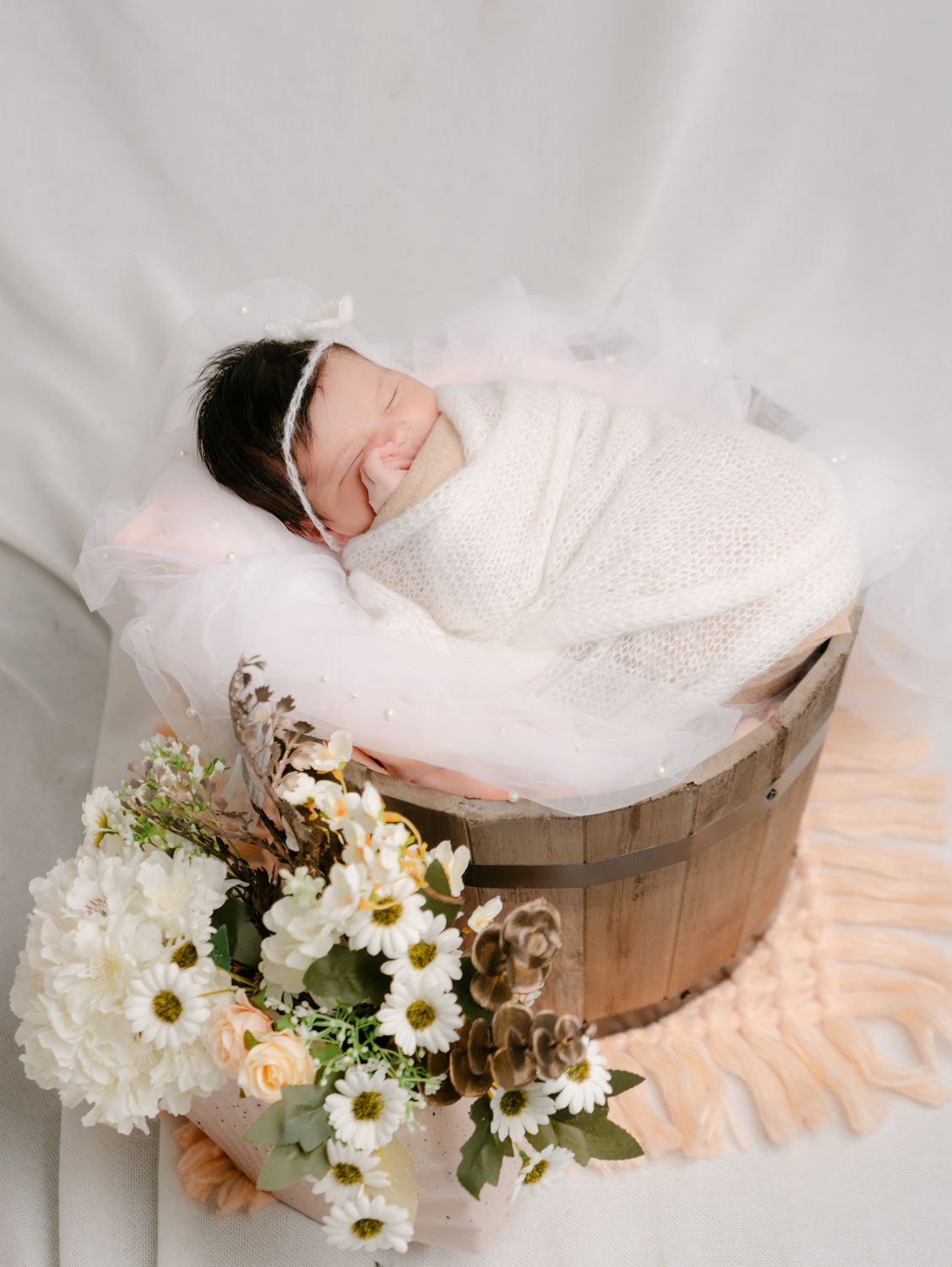 Newborn baby swaddled in white, sleeping in a wooden barrel with white and peach flowers nearby.