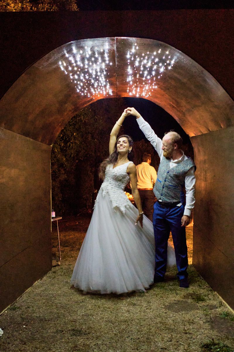 Bride in white gown and groom dancing under glowing arch with sparkling fireworks at night wedding celebration.