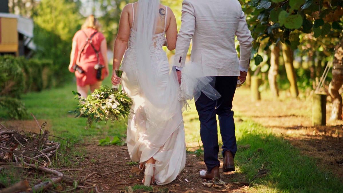 Bride and groom walking hand in hand through vineyard, bride holding bouquet in elegant white wedding dress.