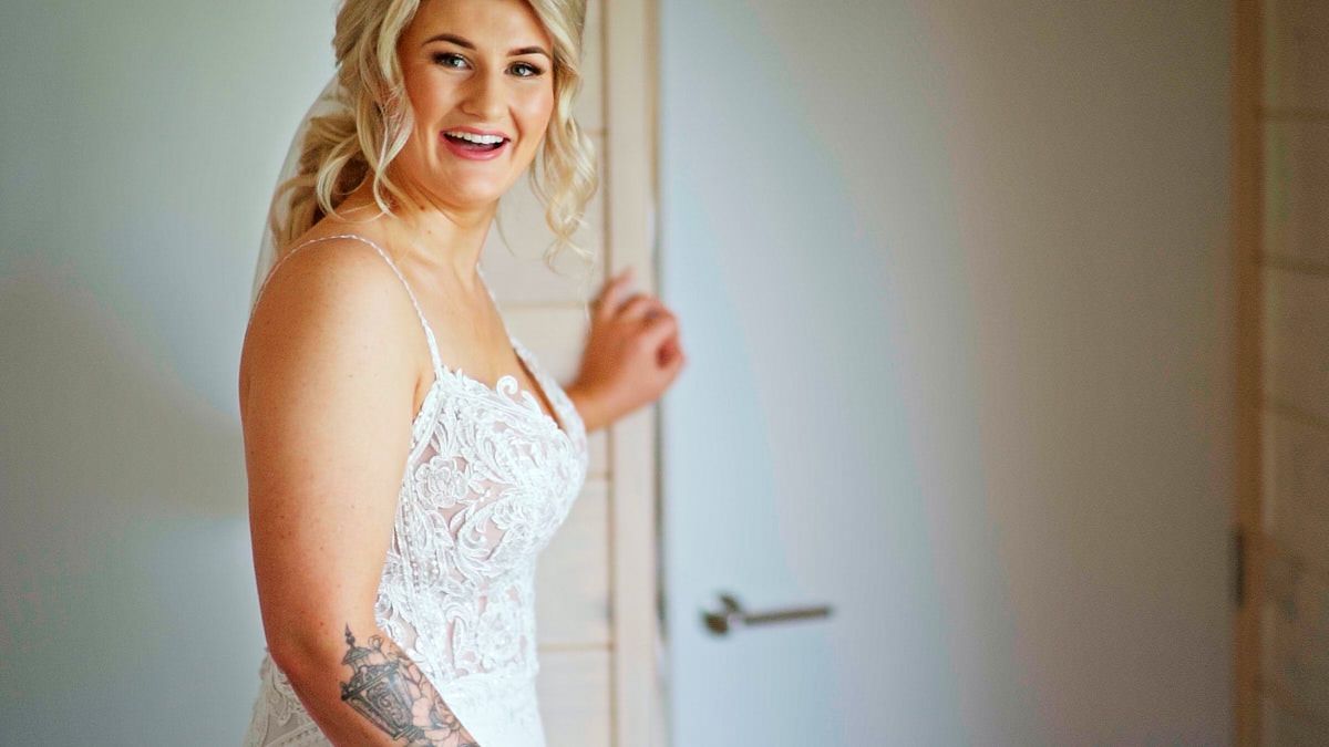Smiling bride in a white lace wedding gown with a tattoo, posing elegantly by a door in natural light.