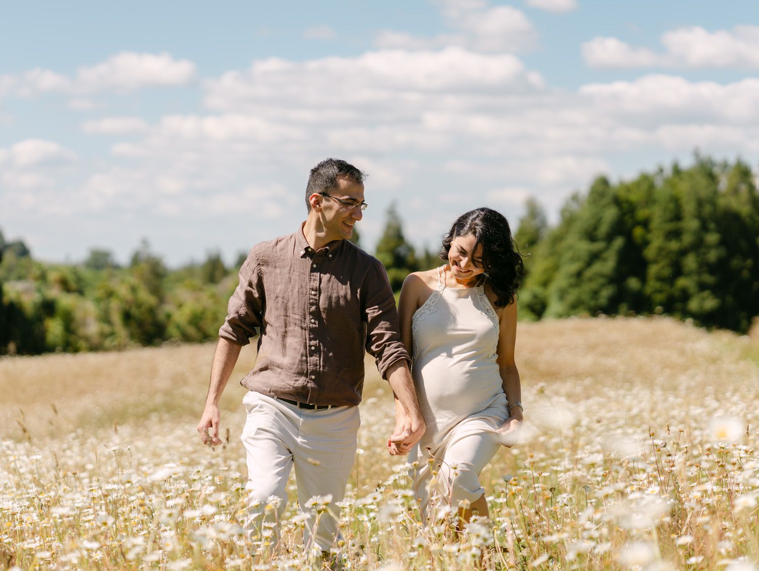 Couple walking hand-in-hand through a sunny wildflower field, woman in white dress showing baby bump.