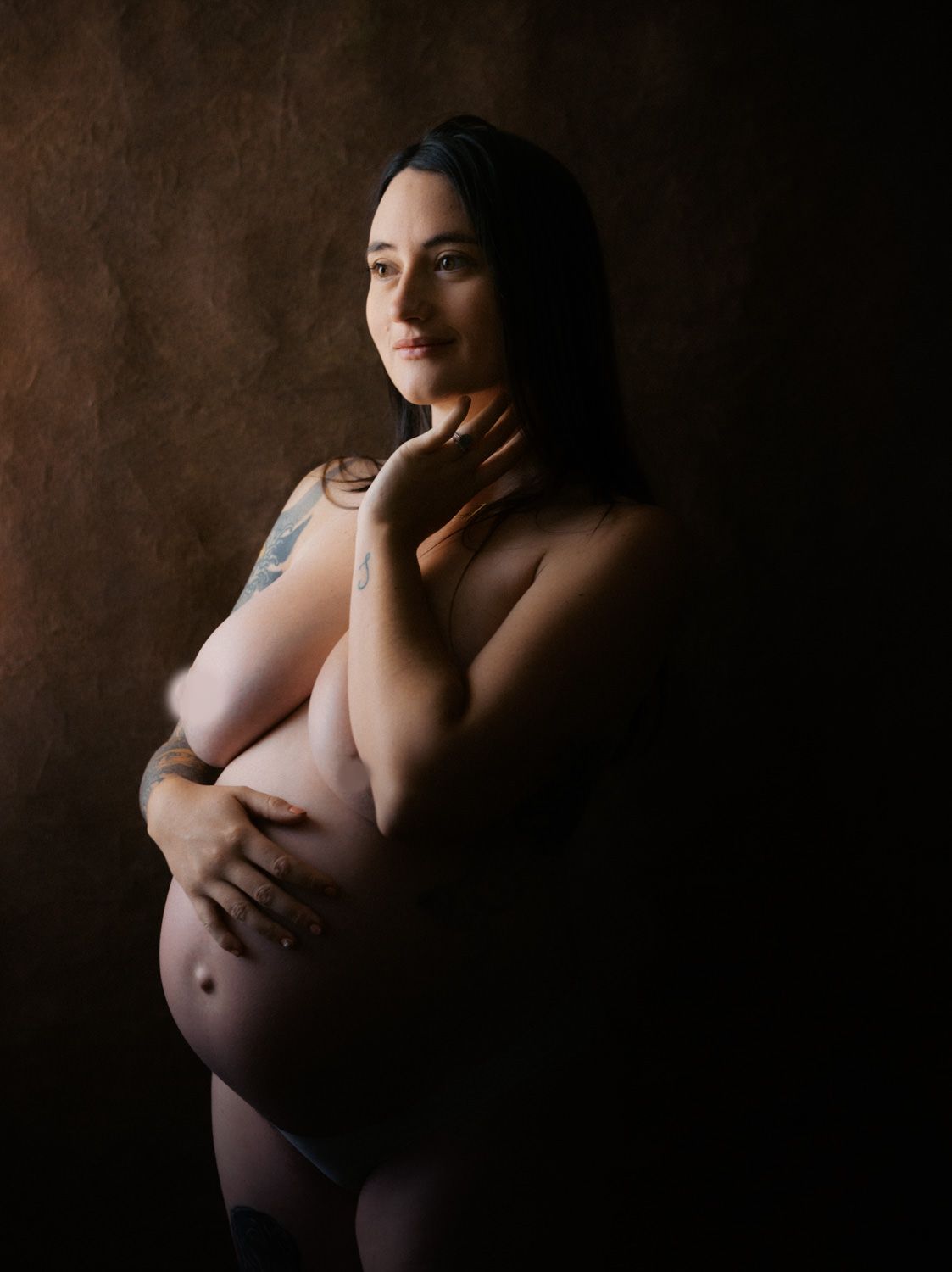 Pregnant woman posing elegantly in a dark studio, cradling her baby bump in a artistic maternity portrait.